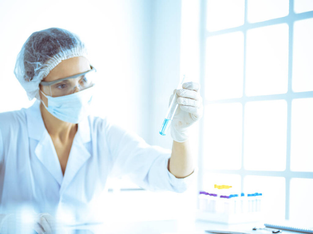 Professional female scientist in protective eyeglasses researching tube with reagents in laboratory toned in blue. Concepts of medicine and science researching - Foto, afbeelding