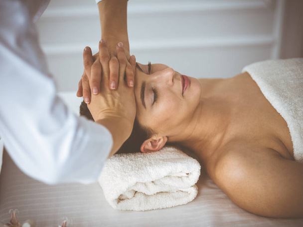 Beautiful brunette woman enjoying facial massage with closed eyes comfortable and blissful. Relaxing treatment in medicine and spa center concepts - Photo, image