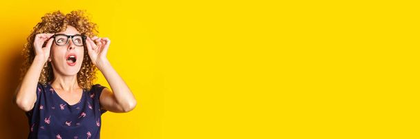 curly young woman looks up in surprise while holding glasses on a yellow background. Banner. - 写真・画像