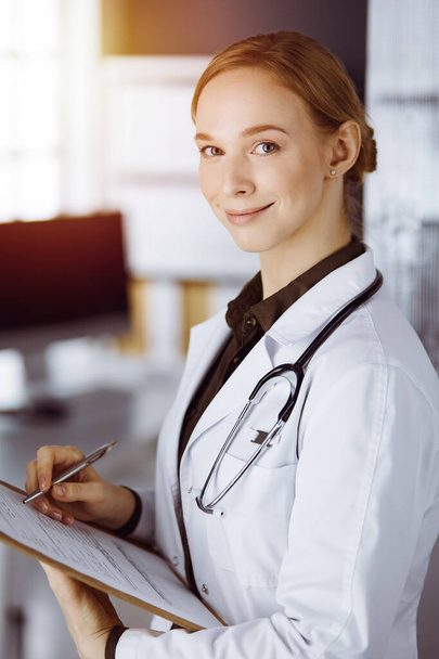 Cheerful smiling female doctor using clipboard in clinic. Portrait of friendly physician woman at work. Medical service in hospital. Medicine concept - Foto, Bild
