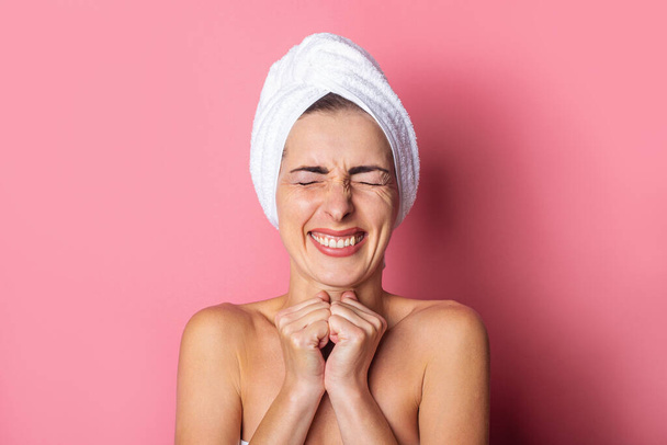 smiling young woman with a towel on her head, eyes tightly closed holds her hands under her chin on a pink background. - Photo, Image