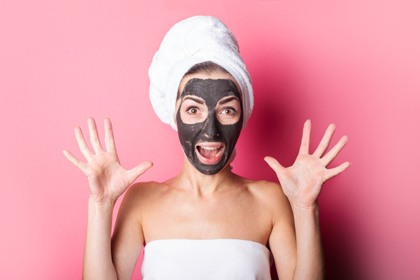 zaskoczona młoda kobieta w masce kosmetycznej na twarzy owinięty ręcznik na głowie na różowym tle. - Zdjęcie, obraz