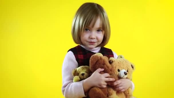 Linda niña rubia con oso de peluche en sus brazos sobre un fondo amarillo - Metraje, vídeo