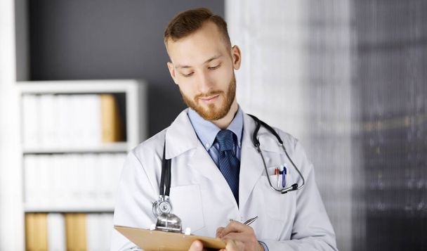 Friendly red-bearded doctor standing and writing with clipboard in clinic at his working place. Perfect medical service in hospital. Medicine, healthcare - Фото, изображение