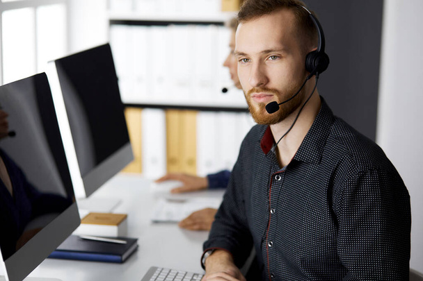 Bearded businessman talking by headset near his female colleague while sitting in modern office. Diverse people group in call center. Telemarketing and customer service - 写真・画像