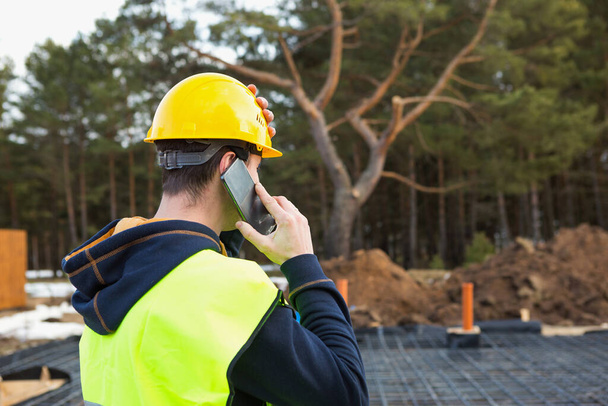 Construction worker talks on a smartphone in a yellow helmet and protective vest against the background of the construction of house-reinforcement for the foundation, pipes and blind area. - Фото, зображення