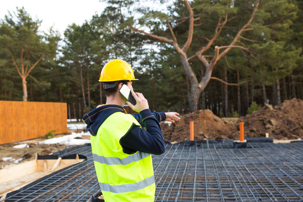 Construction worker talks on a smartphone in a yellow helmet and protective vest against the background of the construction of house-reinforcement for the foundation, pipes and blind area. - Foto, Imagen