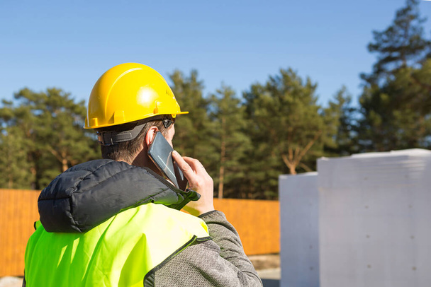 The builder  talking on the phone. Architect in a protective yellow helmet and a signal vest stands backs in front of the construction site of a house with a foundation and aerated concrete blocks.  - Foto, Imagen