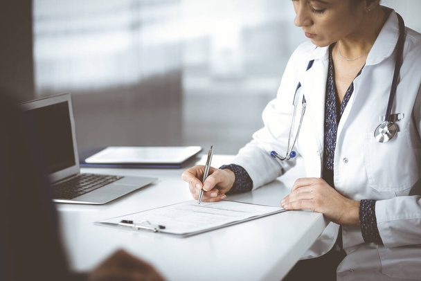 Unknown woman-doctor is writing some medical recommendations to her patient, while they are sitting together at the desk in the cabinet in a clinic. Physician is using a clipboard, close-up. Perfect - Foto, imagen