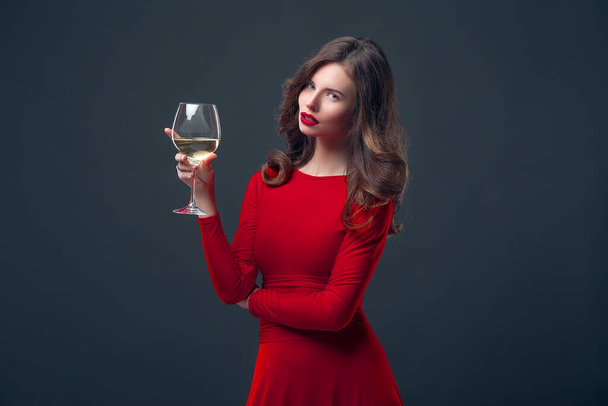 Woman with bright makeup, hairstyle wearing red dress posing with glass of vine over dark background, isolate - Photo, Image