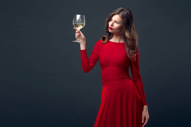Woman with bright makeup, hairstyle wearing red dress posing with glass of vine over dark background, isolate - Photo, Image
