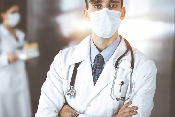 Young male doctor standing with arms crossed in clinic. Smart physician wearing medical protective mask in modern hospital. Medicine and Corona virus concept - Fotó, kép
