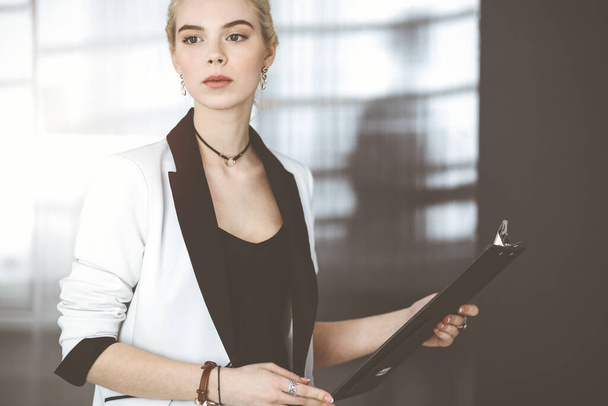 Young blonde business woman or female student in white suit is standing in modern office. Lifestyle and diverse people concept - Фото, изображение