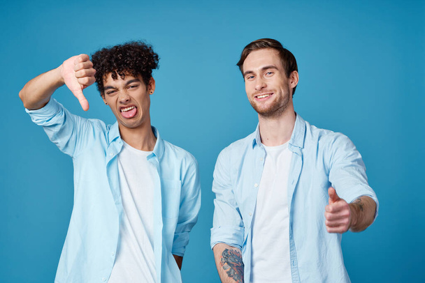 two friends in identical shirts and a t-shirt gesturing with their hands on a blue background - Photo, Image