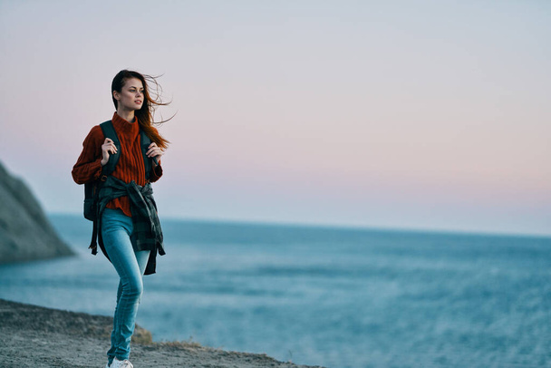 женщина в джинсах с рюкзаком на спине море на заднем плане и высокие горы - Фото, изображение