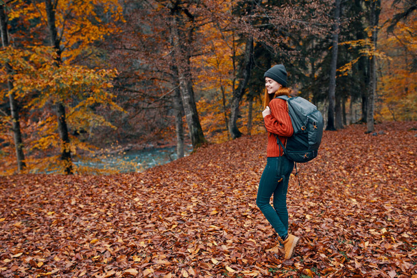 szczęśliwa młoda kobieta z plecakiem w dżinsach buty i sweter chodzą w jesiennym lesie w pobliżu wysokich drzew  - Zdjęcie, obraz