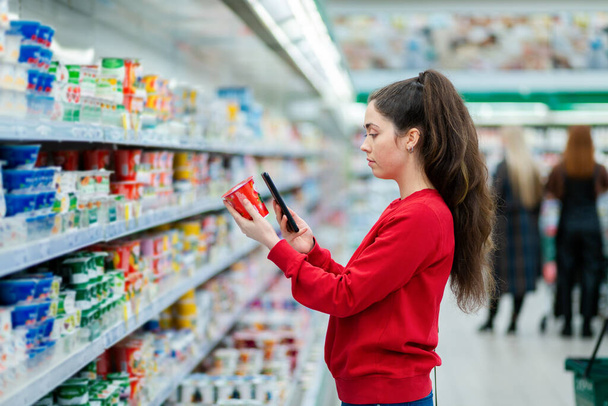 Retrato de mujer joven escanea el código QR en un paquete de yogur. En el fondo, un supermercado con visitantes en un desenfoque. Vista lateral. El concepto de las tecnologías modernas. - Foto, imagen
