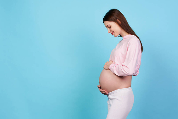 Profile photo, a charming sweet young girl, 9 months pregnant, stands sideways, gently touches her tummy with her hands, smiles, waiting for a meeting with her unborn child. Copy space. - Foto, imagen