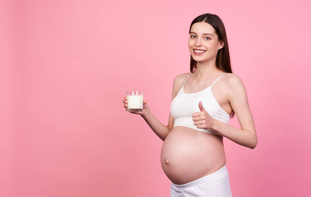 Young attractive Caucasian pregnant woman, brown-haired woman, in a white top and white leggings, drinks milk and shows a like sign on a pink background. Copy space. Pregnancy and nutrition concept. - Fotografie, Obrázek