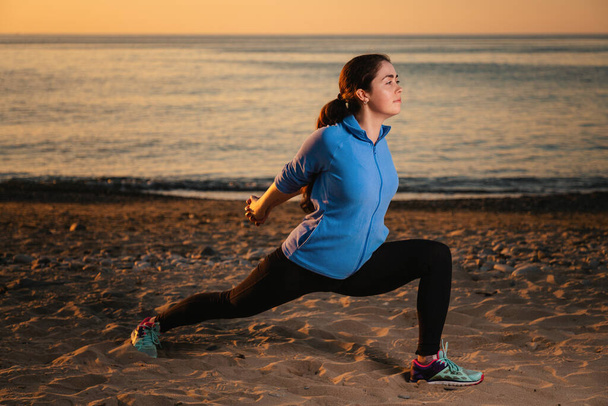 Een jonge vrouw in sportkleding traint door het uitvoeren van een been stretch. Op de achtergrond, zonsondergang, zandstrand en oceaan. Zijaanzicht. - Foto, afbeelding