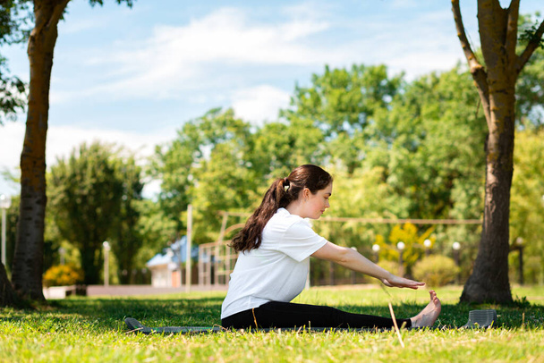 Jóga. Egy fiatal nő sportruhában, egy lábtörlőn ülve, egy parkban a füvön. Oldalnézetből. A sport koncepciója az egészséges életmódért. - Fotó, kép