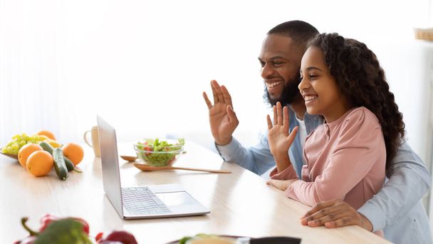 Black man and girl having videocall using laptop waving hello - 写真・画像