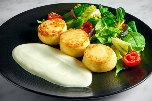 Salted cottage cheese pancakes with sour cream, baked cherry tomatoes and salad, served in a black plate on a marble background. Delicious and healthy breakfast. Ukrainian syrniki - Zdjęcie, obraz