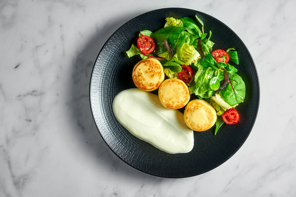 Salted cottage cheese pancakes with sour cream, baked cherry tomatoes and salad, served in a black plate on a marble background. Delicious and healthy breakfast. Ukrainian syrniki - Foto, Bild