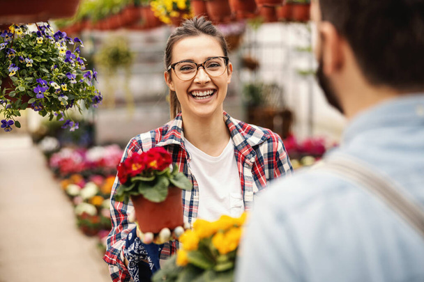 Νεαρή γυναίκα κηπουρός που γελάει στέκεται με το συνάδελφό της και κρατάει την κατσαρόλα με το κόκκινο λουλούδι ενώ κοιτάζει την κάμερα. Επιλεκτική εστίαση στη γυναίκα. - Φωτογραφία, εικόνα
