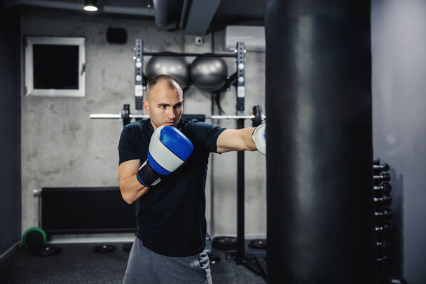 Boxing training. Male boxer hitting a black punching bag at a fitness studio. Male adult boxer wearing gloves and training hard indoor gym. Fitness goal, motivation on training, kicking bag - 写真・画像