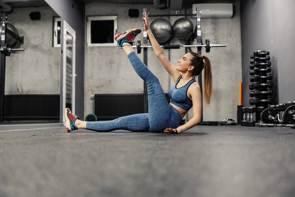 Sports exercises, cross-training abs and legs exercises. Young and beautiful female practices her abdomen and touches her feet with his hands. She wears sports clothes in an indoor gray sports studio - Photo, Image