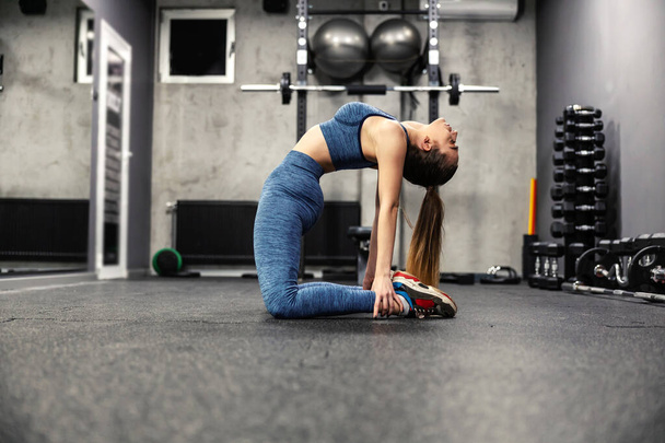 Stretching en uitbreiding van de spieren van het lichaam. Een elastische vrouw in goede fysieke vorm strekt de spieren van het lichaam in de sportschool. Ze strekt haar buikspieren uit door haar handen op haar benen te leggen. - Foto, afbeelding