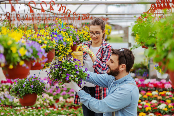 Couple in love standing in greenhouse and taking care of flowers. Man holding pot while woman watering flowers. - Photo, Image
