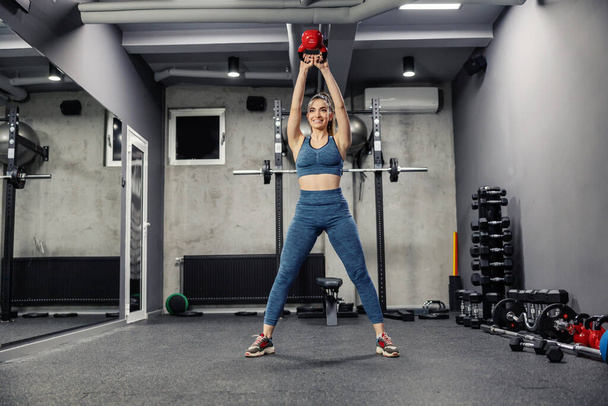 Swing exercising with a kettle bell to pump the whole body muscles. A front view of a sexy woman in sportswear and in good physical shape lifting weights in an indoor gym. Fitness challenge - Photo, Image