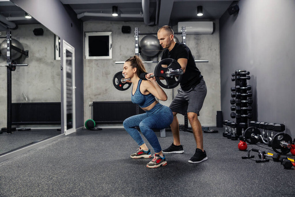 Barbell squats with the help of personal training. Fit woman in sportswear and in good shape does barbell squats to strengthen the muscles of the whole body. Coach assistance in individual training - Photo, Image