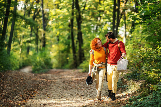 Ein junges glückliches Paar, das sich umarmt und in der Natur spazieren geht. Paar hält Picknickutensilien in der Hand. - Foto, Bild