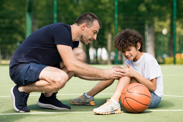 Profesor ayudando a niño con trauma de rodilla después de jugar baloncesto - Foto, Imagen