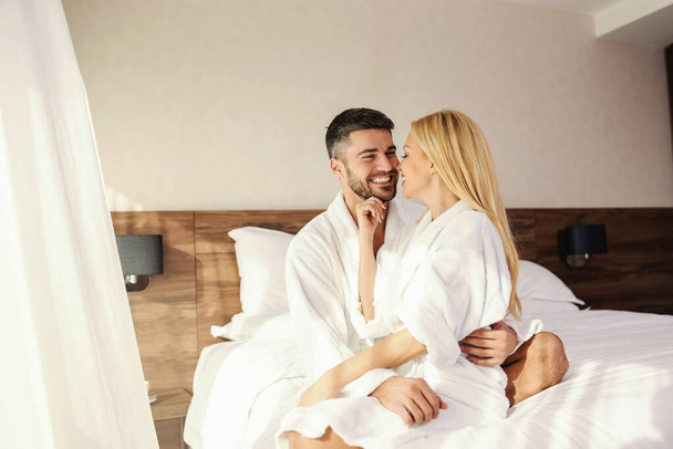 Onnellinen pari halailemassa sängyssä hotellihuoneessa. Romanttiset aamut hotellissa. Mies ja nainen valkoisissa vaatteissa ja lämpimän hotellihuoneen valkoisissa lakanoissa halailevat toisiaan. Rakkausmuistot - Valokuva, kuva