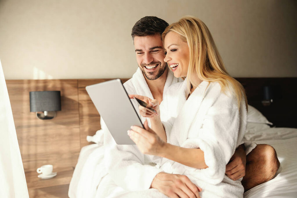 The young couple travels together in their free time. A beautiful blonde is sitting in the lap of a handsome man. They are happy and excited and use a tablet and a credit card for online reservation - Photo, Image