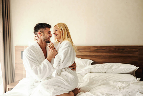 Kameralne poranne chwile w hotelowym łóżku w cieple szlafroka. Romantyczne ujęcie dwojga kochanków, kobiety w ramionach mężczyzny trzymającego ją za rękę. Mężczyzna i kobieta wyglądają na szczęśliwych i spełnionych. - Zdjęcie, obraz