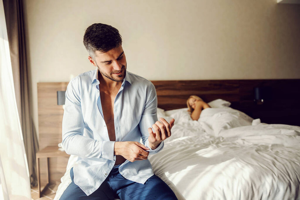Επιθυμία και πάθος σε ένα δωμάτιο ξενοδοχείου. Το πρωί μετά τον έρωτα και το βρώμικο άσπρο κρεβάτι. Ο άντρας κουμπώνει ένα κουμπί στο μανίκι της μπλούζας του ενώ η γυναίκα τον παρακολουθεί. Αγάπη, πάθος, σεξ - Φωτογραφία, εικόνα