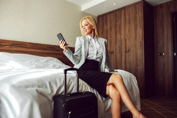 Einde van een zakenreis. Mooie en succesvolle vrouw in formele kleding zit op het bed in een hotelkamer en gebruikt een telefoon. Naast haar staat een zwarte koffer die ze klaarmaakt om uit te checken in het hotel. - Foto, afbeelding