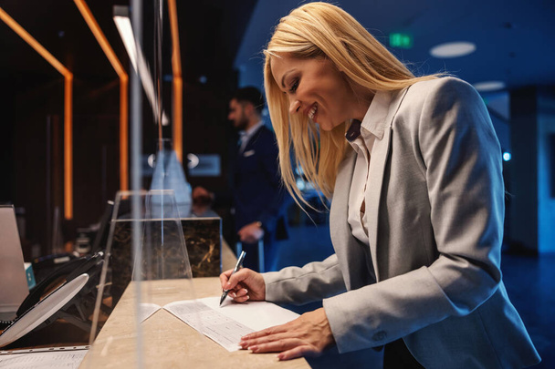 Happy businesswoman standing at reception and checking in. She is filling up the form. Business trip, luxurious hotel, weekend - Foto, Imagem