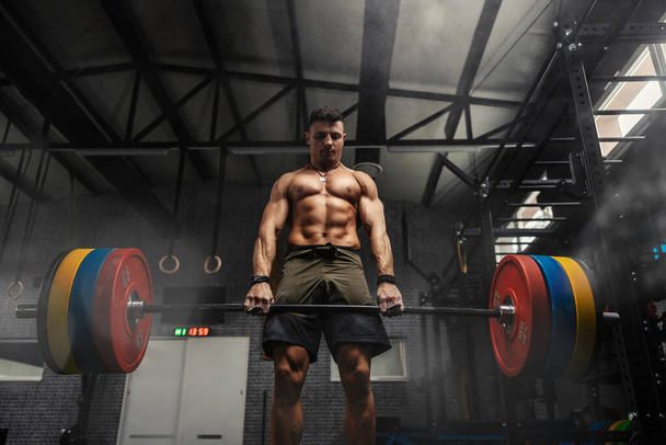 Weightlifter at the gym, a moment before a powerful movement. A man with a strong body holds a heavy barbell in his hands and does a dead lift in a dark atmosphere gym. Motivation in sports, cross fit - Photo, Image