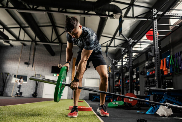 Preparation of weights with a bar for training strong muscle burning. A man in sportswear sets weights with a barbell in the gym. Cross fit and boot camp training - Photo, Image
