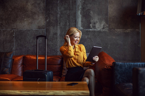 Positive businesswoman sitting in a hall of a hotel and checking in a hotel online. She is on a business trip. Telecommunications, travel, business trip - Zdjęcie, obraz