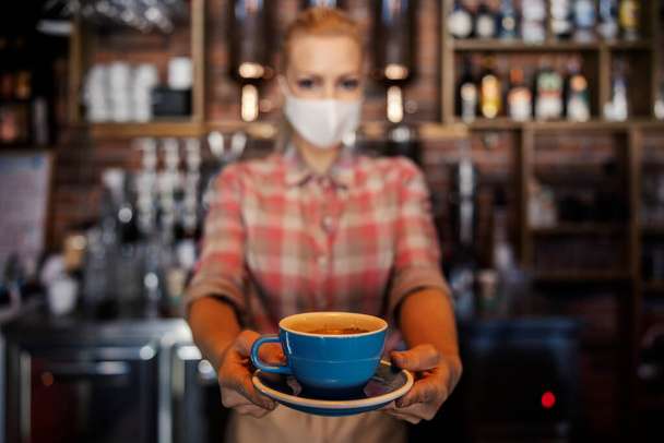 The waitress offered a cup of coffee. A photo of an isolated blue cup of coffee behind a blurred background of a woman with a protective face mask. Female hands hold the cup and extend it to the guest - Photo, Image