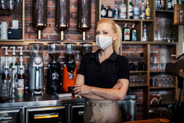 Trinkservice während der Coronavirus-Pandemie. Die Frau steht an der Theke des Restaurants und trägt ein schwarzes T-Shirt und eine Schürze. Eine Kellnerin hat die Arme über einer Gesichtsschutzmaske verschränkt - Foto, Bild