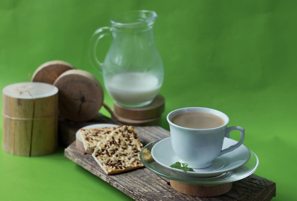手前の白いカップには、木の板の上にココアドリンクと緑の葉、種子とパンの背景の愛と木製のコースターに牛乳とデカンタです。 - 写真・画像