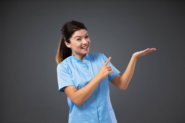 Ευχάριστη και χαμογελαστή νοσοκόμα. Πορτρέτο μιας νεαρής και όμορφης νοσοκόμας με μπλε στολή που στέκεται μπροστά σε γκρι φόντο και δείχνει την κατεύθυνση της κίνησης με τα χέρια της - Φωτογραφία, εικόνα
